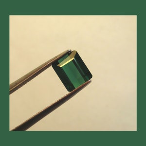 May include: A deep green emerald gemstone held in tweezers. The emerald is cut into a rectangular shape with beveled edges.