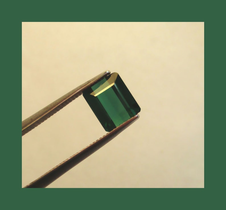 May include: A deep green emerald gemstone held in tweezers. The emerald is cut into a rectangular shape with beveled edges.