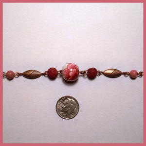 May include: A copper chain bracelet with red and pink beads, including a white porcelain bead with a pink floral design. The bracelet is laid out on a white surface with a US dime for scale.