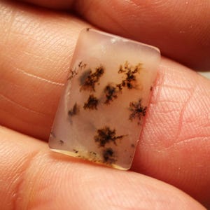 May include: A rectangular, polished gemstone with a translucent, pale pink base. The stone features intricate, tree-like patterns in shades of brown and black, creating a natural, organic design. The gemstone is held in a hand.