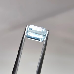 May include: A rectangular, light blue gemstone held by a pair of tweezers. The gem has a clear, transparent appearance with visible internal facets. The tweezers are dark gray, contrasting with the bright gem.