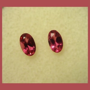 May include: Two oval-shaped pink gemstones, likely rubies, set against a beige background.