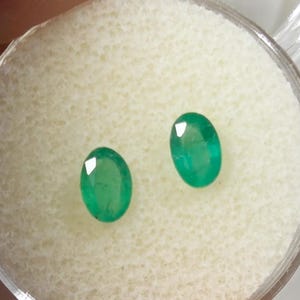 May include: Two oval-cut emerald gemstones, each with a vibrant green hue and a faceted surface. The gems are displayed in a clear, round container with a white, textured interior, showcasing their brilliance.