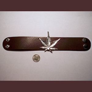 May include: A brown leather bracelet with a silver marijuana leaf charm. The bracelet has two silver snaps on the side.