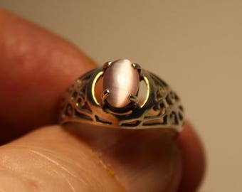 May include: A silver ring with an oval, light pink gemstone. The ring has an open filigree design around the band. The gemstone is set in a gold-colored bezel.