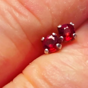 May include: A pair of small stud earrings featuring round, faceted, deep red gemstones set in four-prong silver-toned metal settings. The stones exhibit a vibrant crimson hue with clear internal reflections and a polished, reflective surface finish.
