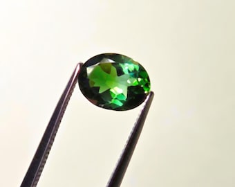 May include: An oval-cut, vibrant green gemstone is held by a pair of silver tweezers. The gem has multiple facets that reflect light, creating a sparkling effect. The background is a soft, neutral color.
