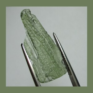 May include: A translucent green crystal with a striated texture, held by tweezers. The crystal is a natural formation with a unique, almost wood-like appearance.