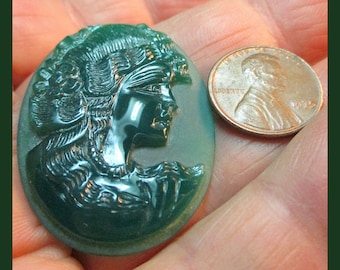 May include: A green cameo carved with a detailed profile of a woman's face. The cameo is oval shaped and is about the size of a US penny.