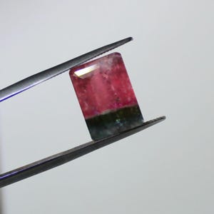 May include: A rectangular gemstone, held by tweezers, displays a gradient of colors. The top is a translucent white, transitioning to a vibrant pink, and ending with a dark green base. The gem is likely a tourmaline.