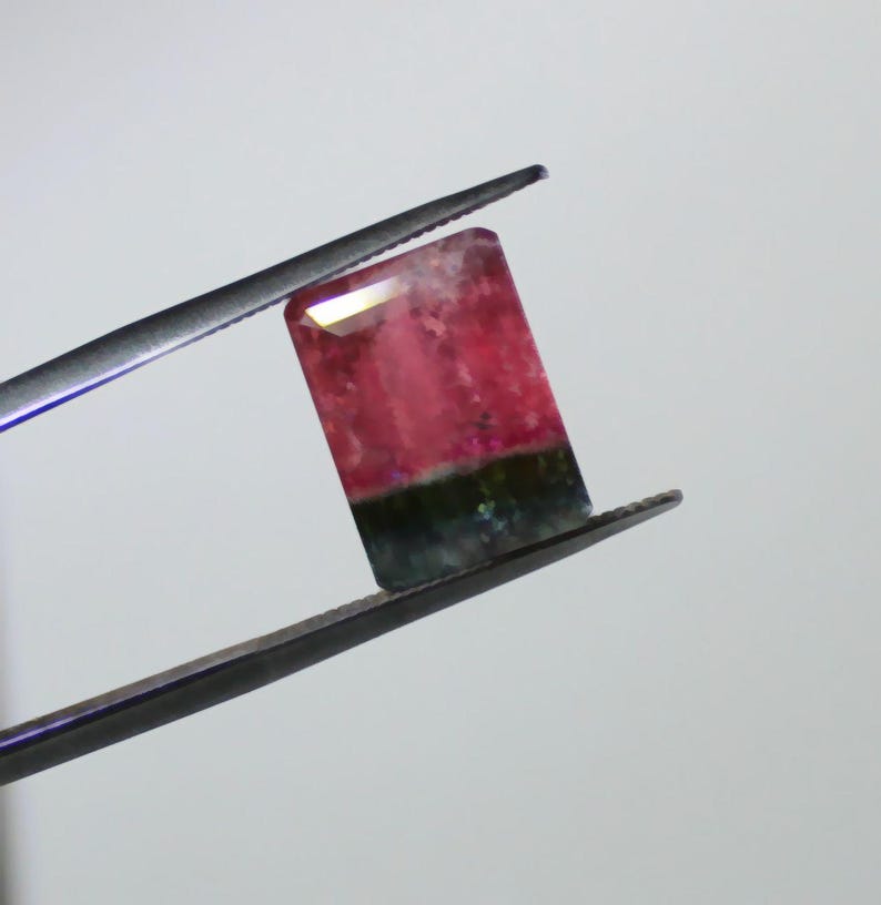 May include: A rectangular gemstone, held by tweezers, displays a gradient of colors. The top is a translucent white, transitioning to a vibrant pink, and ending with a dark green base. The gem is likely a tourmaline.