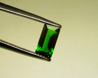 May include: A vibrant green gemstone, likely a peridot, is held in tweezers. The gemstone is rectangular in shape with a faceted cut.