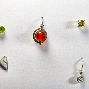 May include: A collection of jewelry pieces including a round green gemstone stud, a spherical orange bead pendant in a silver frame, a yellow gemstone stud, a triangular opal-like stud, and a small clear gemstone drop earring with a silver hook.