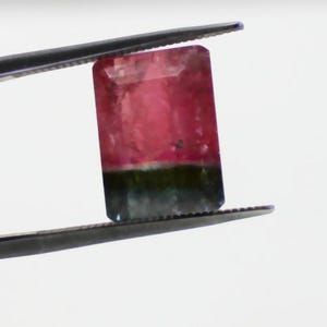 May include: A rectangular watermelon tourmaline gemstone, held by tweezers. The gem features a gradient of colors, with a pink top, a green base, and a clear, faceted surface. The tweezers are dark gray.