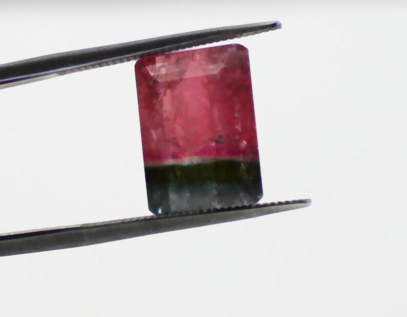 May include: A rectangular watermelon tourmaline gemstone, held by tweezers. The gem features a gradient of colors, with a pink top, a green base, and a clear, faceted surface. The tweezers are dark gray.