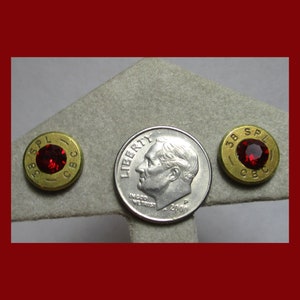 May include: Two gold-colored bullet casings with red gemstones set into the center. The casings are marked "38 SPL CBC". A US dime is included for size comparison.