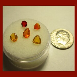May include: A collection of faceted gemstones in various shapes and shades of orange and yellow, displayed in a clear plastic container. A U.S. dime is placed next to the container for size comparison. The gemstones include round, pear, and triangle shapes.