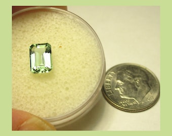 May include: A light green emerald cut gemstone in a small plastic container next to a US dime coin.