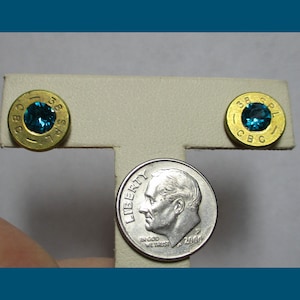 May include: A pair of earrings made from gold-colored bullet casings with blue gemstones set in the center. The casings are marked "38 SPL CBC" and "58 SPL CBC".
