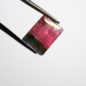 May include: A rectangular gemstone with a watermelon color scheme, held by tweezers. The gem features a vibrant pink center transitioning to a dark green base. The edges are faceted, catching the light.