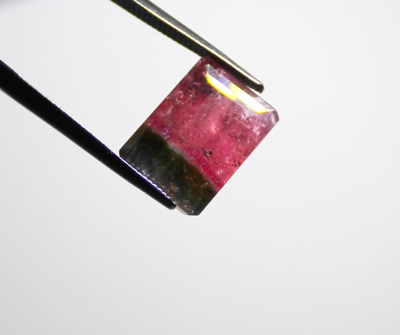 May include: A rectangular gemstone with a watermelon color scheme, held by tweezers. The gem features a vibrant pink center transitioning to a dark green base. The edges are faceted, catching the light.