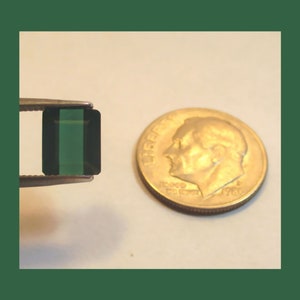 May include: A dark green emerald gemstone in a rectangular cut, set in a silver bezel setting. The gemstone is approximately the size of a dime.