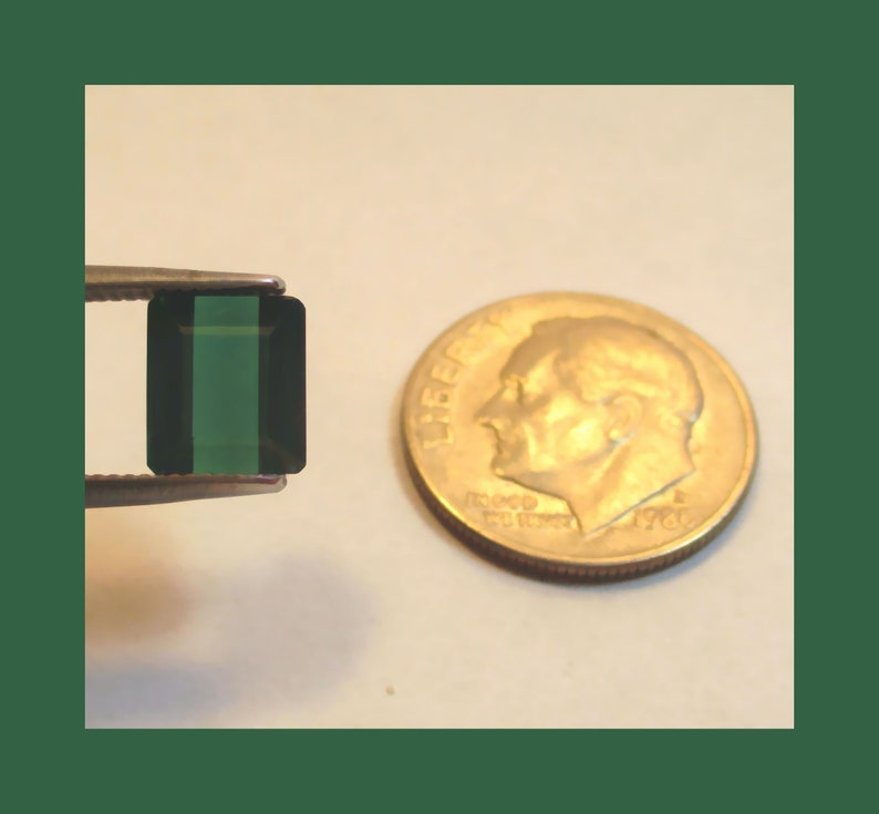 May include: A dark green emerald gemstone in a rectangular cut, set in a silver bezel setting. The gemstone is approximately the size of a dime.