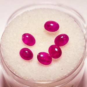 May include: A collection of six oval-shaped, vibrant red gemstones. The polished stones are displayed in a clear, round container with a white, textured interior. The gems have a high luster and are evenly spaced.