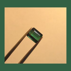 May include: A dark green gemstone with a rectangular shape is held by tweezers against a light beige background.
