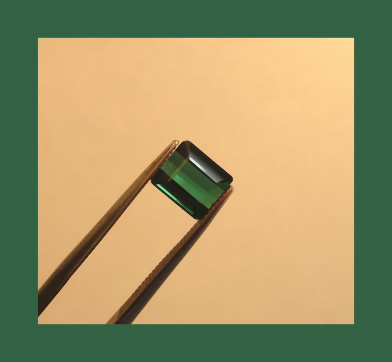 May include: A dark green gemstone with a rectangular shape is held by tweezers against a light beige background.