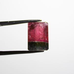 May include: A rectangular gemstone held by tweezers. The gem features a gradient of colors, with a vibrant pink hue at the top transitioning to a darker green at the bottom. The gem is set against a plain white background.