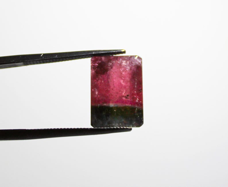 May include: A rectangular gemstone held by tweezers. The gem features a gradient of colors, with a vibrant pink hue at the top transitioning to a darker green at the bottom. The gem is set against a plain white background.