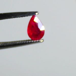 May include: A vibrant, teardrop-shaped ruby gemstone held by tweezers. The ruby displays a deep red hue with facets catching the light. The gemstone is the focal point against a neutral background, showcasing its clarity and color.