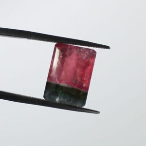 May include: A rectangular gemstone with a vibrant color gradient, held by tweezers. The top half is a deep pink, transitioning to a dark green at the base. The gem's facets catch the light, showcasing its polished surface.