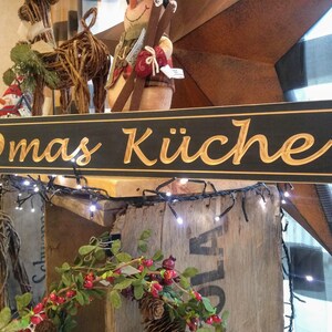 May include: A black wooden sign with gold lettering that reads "Omas K&uuml;che". The sign is hanging above a wooden shelf with a string of white lights.