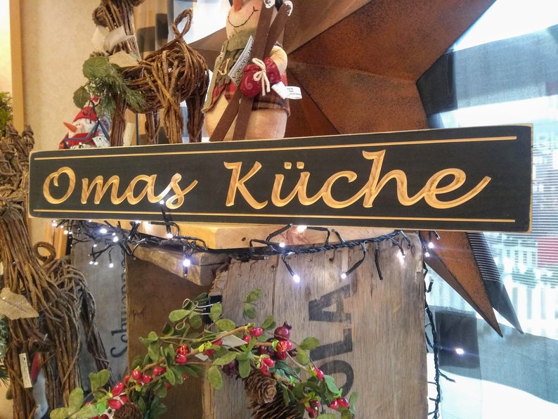 May include: A black wooden sign with gold lettering that reads "Omas K&uuml;che". The sign is hanging above a wooden shelf with a string of white lights.