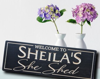 May include: Black wooden sign with white text that reads "Welcome to Sheila's She Shed". The sign is sitting on a light blue surface in front of two white vases with purple flowers.