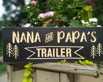 May include: A black wooden sign with gold lettering that reads "Nana and Papa's Trailer". The sign has a stylized banner and pine tree graphics.