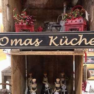 May include: A black wooden sign with gold lettering that reads "Omas K&uuml;che". The sign is hanging on a wooden shelf with other decorative items.