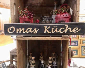 Puede incluir: Un letrero de madera negro con letras doradas que dice "Omas Küche". El letrero está colgado en un estante de madera con otros artículos decorativos.