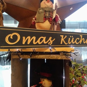 May include: A black wooden sign with gold lettering that reads "Omas K&uuml;che". The sign is hanging above a wooden crate with a snowman figurine inside.