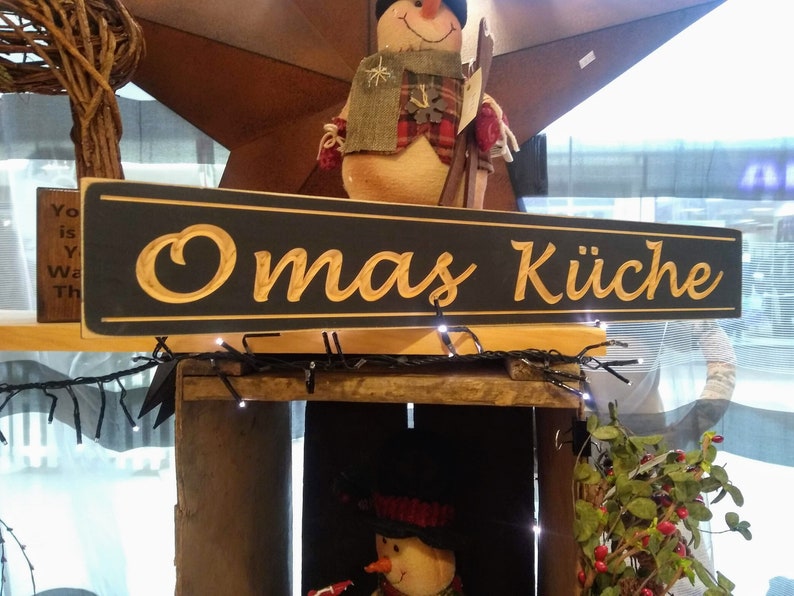 May include: A black wooden sign with gold lettering that reads "Omas K&uuml;che". The sign is hanging above a wooden crate with a snowman figurine inside.