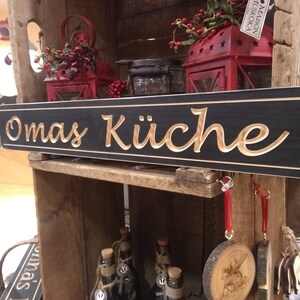 May include: A black wooden sign with the words "Omas K&uuml;che" carved in gold lettering. The sign is hanging on a wooden shelf with other decorative items.