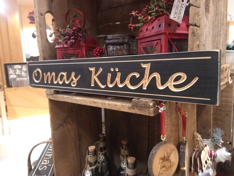 May include: A black wooden sign with the words "Omas K&uuml;che" carved in gold lettering. The sign is hanging on a wooden shelf with other decorative items.