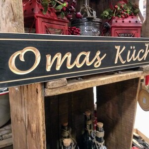 May include: A black wooden sign with the words "Omas K&uuml;che" carved in a light wood color. The sign is hanging on a wooden shelf with other decorative items.