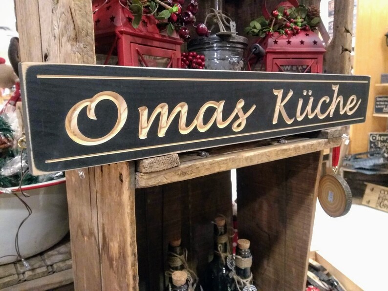 May include: A black wooden sign with the words "Omas K&uuml;che" carved in a light wood color. The sign is hanging on a wooden shelf with other decorative items.