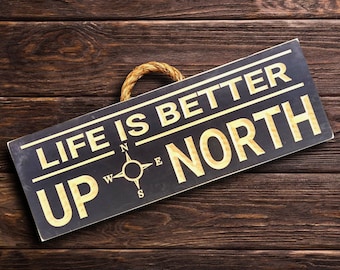 May include: Black wooden sign with white rope hanging from the top. The sign reads "Life is better up north" with a compass graphic in the center.
