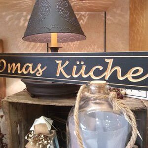May include: A black wooden sign with gold lettering that reads "Omas K&uuml;che". The sign is hanging above a wooden crate and a glass jar with a rope handle.