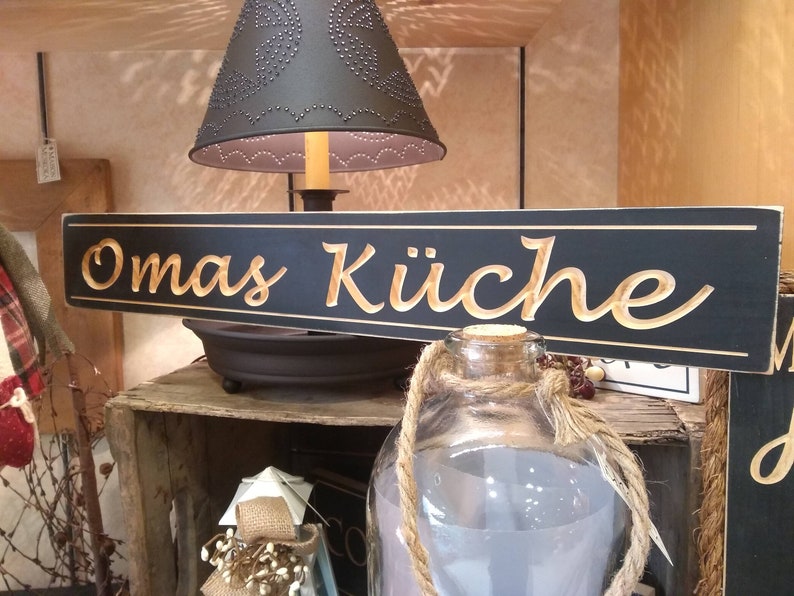 May include: A black wooden sign with gold lettering that reads "Omas K&uuml;che". The sign is hanging above a wooden crate and a glass jar with a rope handle.