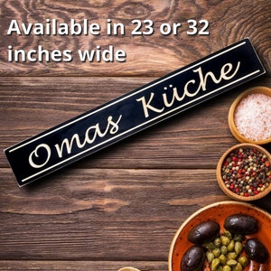 May include: A black wooden sign with gold lettering that reads "Omas K&uuml;che" on a rustic wooden background. The sign is available in 23 or 32 inches wide.